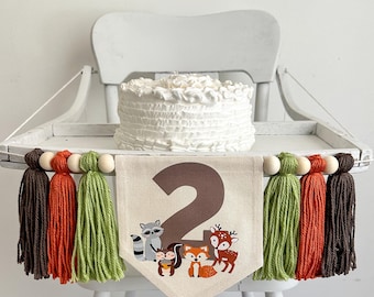 Puede incluir: Una silla alta blanca con un pastel blanco en una bandeja. La bandeja tiene una pancarta con el número "2" y animales del bosque. La pancarta está decorada con borlas de hilo marrón, naranja y verde.