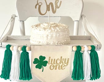 May include: White high chair with a white cake on a tray. The cake is decorated with white frosting. A green and white tassel garland hangs from the tray. A banner with a green shamrock and gold glitter text that reads "lucky one" hangs from the tray. A gold glitter "one" topper sits on top of the cake.