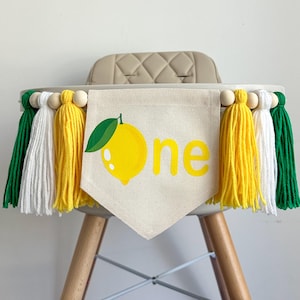 May include: A high chair decorated with a lemon-themed banner that says "one" and yellow, white, and green tassel garland.