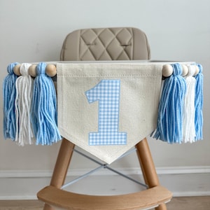 May include: A high chair banner with a light beige fabric base and a blue gingham number one applique. The banner is decorated with blue, white, and wooden bead tassels. The high chair is made of wood and has a beige seat.