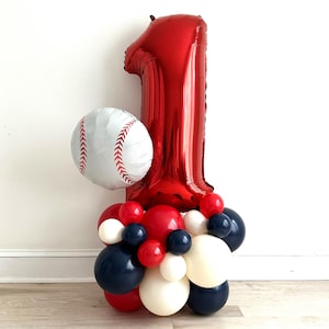 May include: A baseball-themed balloon arrangement for a first birthday. It features a large red number one balloon, a baseball-shaped balloon, and a cluster of red, white, and navy blue balloons at the base. The balloons are arranged on a light-colored surface.