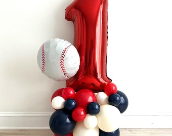 Pode incluir: Um arranjo de balões com tema de beisebol para um primeiro aniversário. Apresenta um grande balão vermelho com o número um, um balão em forma de bola de beisebol e um grupo de balões vermelhos, brancos e azul marinho. Os balões estão dispostos em uma superfície clara.