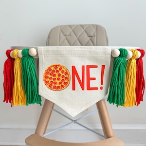 Op de afbeelding: Een slinger voor een kinderstoel met een pizza-afbeelding en het woord "ONE!" in het rood. De slinger is versierd met rode, gele en groene kwastjes en houten kralen. De slinger is bevestigd aan een houten kinderstoel.