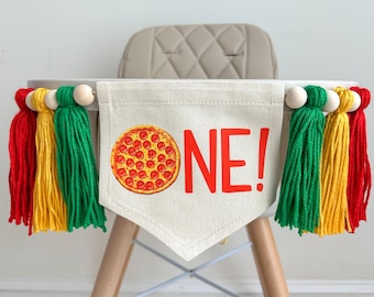 May include: A high chair banner with a pizza graphic and the word "ONE!" in red. The banner is decorated with red, yellow, and green tassels and wooden beads. The banner is attached to a wooden high chair.