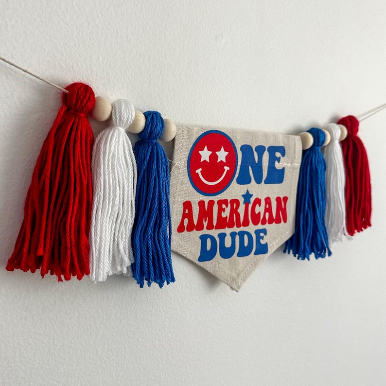 May include: A red, white and blue tassel garland with a fabric banner that reads "ONE AMERICAN DUDE" with a smiley face and stars.
