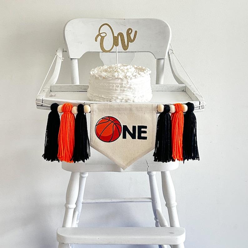 May include: White high chair with a banner that says "ONE" and a basketball graphic. The banner is decorated with black and orange tassels. A white cake with a gold "One" topper sits on the chair.
