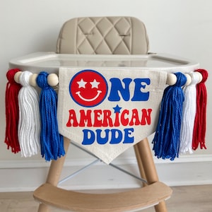 A 5 by 7 inch canvas flag with the words ONE AMERCAN DUDE in red and blue with a red smile face as the O in one. On each side there is a 5 inch royal blue, white and red yarn tassel each separated by a wooded bead for a total of 6 tassels.