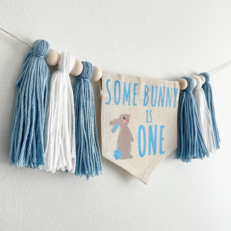 May include: A blue and white tassel banner with the text "Some Bunny is One" and a cartoon bunny illustration. The banner is strung with wooden beads.