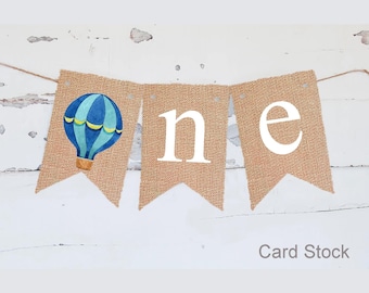 May include: A burlap banner with the word "one" in white letters. The first "o" is replaced with a blue and yellow hot air balloon.