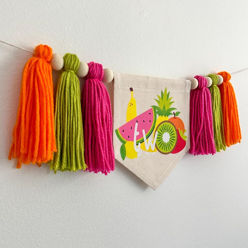 May include: A colorful fruit-themed banner with the word "TWO" printed on it. The banner is decorated with tassels in orange, green, and pink.