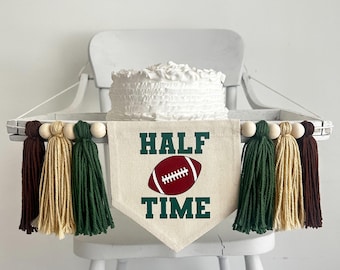 May include: A white high chair with a white cake on top and a green and brown banner that says "HALF TIME" with a red football graphic.