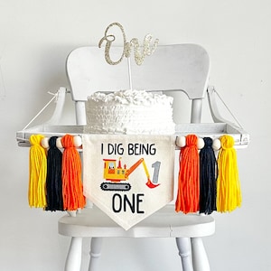 Peut inclure: Une chaise haute blanche avec un plateau blanc et une bannière qui dit "I Dig Being One" avec un dessin d'excavateur jaune. La bannière est décorée de glands jaunes, orange et noirs. Un gâteau blanc avec un topper "One" en paillettes dorées est posé sur le plateau.