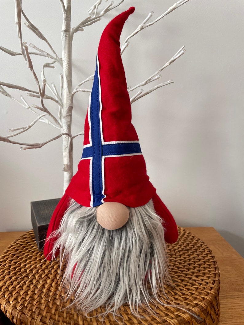 May include: A red and blue gnome with a white beard wearing a hat with the Norwegian flag on it.