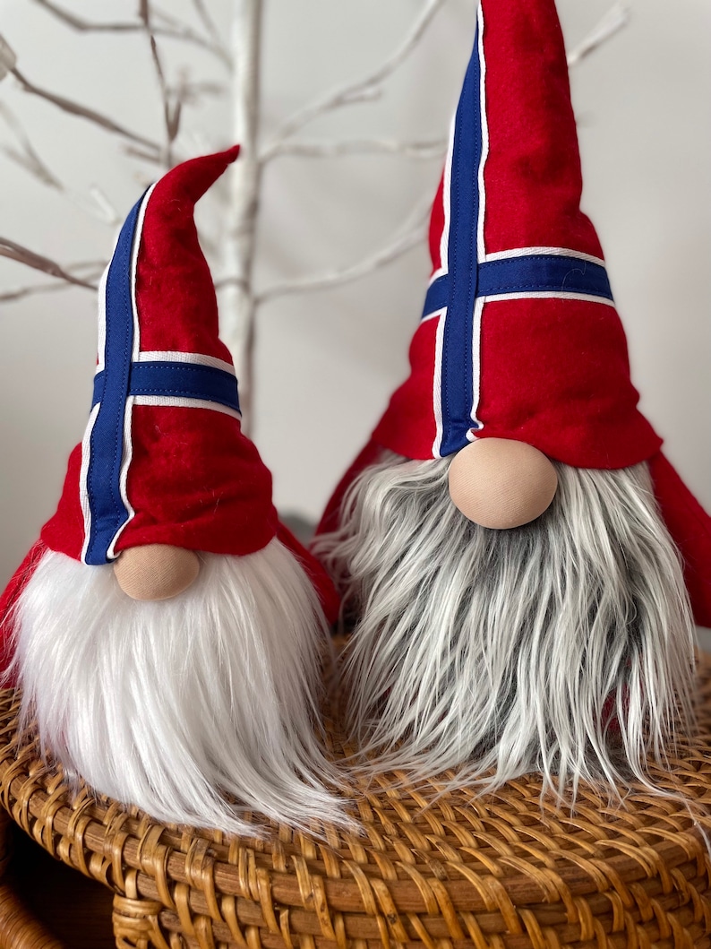 May include: Two gnomes with white beards and red hats decorated with the Norwegian flag. The gnomes are sitting on a woven wicker basket.