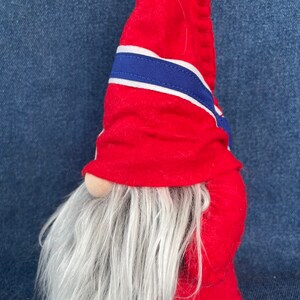 May include: A red felt gnome with a white and blue striped hat and a long white beard. The gnome is standing on a blue denim background.