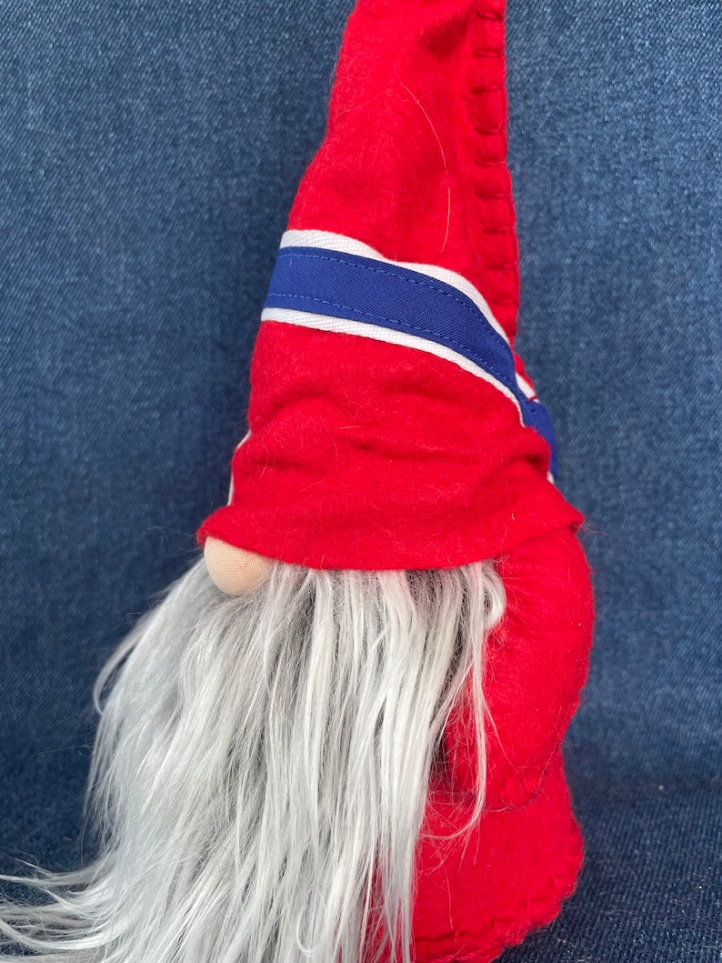May include: A red felt gnome with a white and blue striped hat and a long white beard. The gnome is standing on a blue denim background.