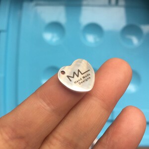 Puede incluir: Encanto de coraz&oacute;n plateado con un logotipo grabado en negro que dice "ML Hand Made Designs".