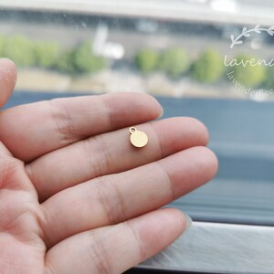 Peut inclure: Un petit charme rond en m&eacute;tal dor&eacute; avec une boucle en haut. Le charme est tenu dans la main d'une personne.