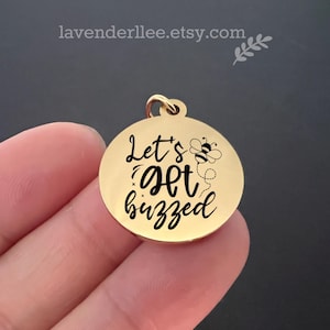 Könnte beinhalten: Goldfarbener Metallanhänger mit einer schwarzen Aufschrift "Let's get buzzed" und einer kleinen schwarz-weißen Bienenillustration.