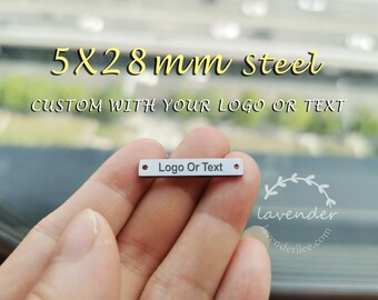 Op de afbeelding: Een close-up van een zilveren metalen label met afgeronde randen. Het label is 5 mm breed en 28 mm lang. Het label heeft een gat aan elk uiteinde en de tekst "Logo Or Text" is op het label gedrukt.