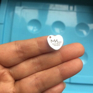 Puede incluir: Un charm de plata en forma de coraz&oacute;n con las letras "ML" y el texto "Hand Made Designs" grabado en &eacute;l.