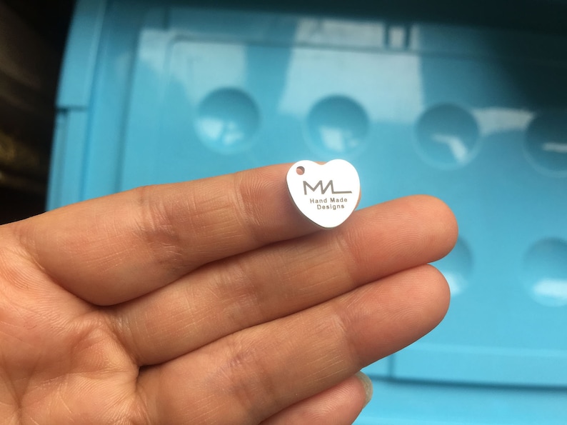 Puede incluir: Un charm de plata en forma de coraz&oacute;n con las letras "ML" y el texto "Hand Made Designs" grabado en &eacute;l.