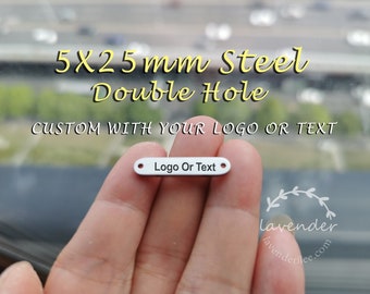 Op de afbeelding: Een kleine, rechthoekige, zilverkleurige metalen tag met twee gaten. De tag leest "Logo Or Text" en is gelabeld "5X25mm Steel Double Hole". Ontworpen om te worden aangepast met een logo of tekst.