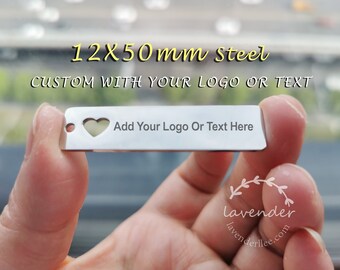 Op de afbeelding: Een rechthoekig stalen label van 12 mm x 50 mm, met een hartvormige uitsparing en een gat. Het label leest "Add Your Logo Or Text Here" en "CUSTOM WITH YOUR LOGO OR TEXT". Het label wordt door een hand omhoog gehouden.