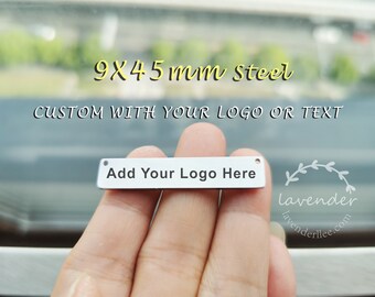 Op de afbeelding: Een klein, rechthoekig stalen label, 9 x 4,5 mm, met de tekst "Add Your Logo Here" in zwart. Het label is ontworpen om te worden aangepast met een logo of tekst. De afbeelding bevat ook de tekst "CUSTOM WITH YOUR LOGO OR TEXT".
