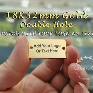 Puede incluir: Una etiqueta rectangular dorada con dos agujeros, que mide 18 x 32 mm. La etiqueta tiene el texto "Add Your Logo Or Text Here" y "CUSTOM WITH YOUR LOGO OR TEXT". La etiqueta se sostiene en una mano.