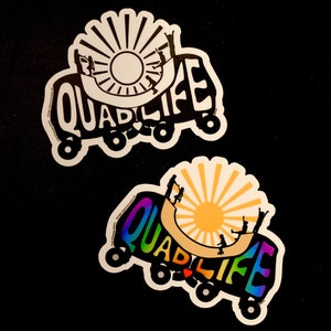 May include: Two stickers with the word "QUADLIFE" in a stylized font, featuring a sun and silhouettes of people roller skating. One sticker is black and white, the other has a rainbow gradient.