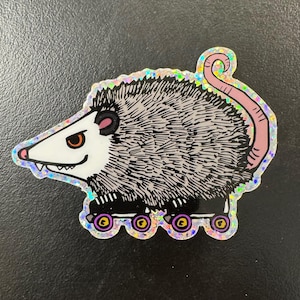 May include: A gray and white opossum wearing roller skates. The opossum has a pink tail and a toothy grin. The roller skates are black with colorful wheels.