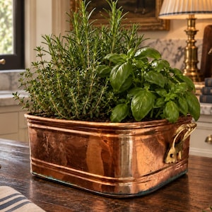 May include: A rectangular copper planter with two side handles containing a lush arrangement of fresh green herbs, including rosemary and basil. The metallic container features a polished finish with visible reflections and a sturdy, wide base.