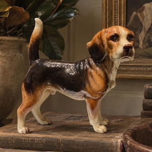 May include: A ceramic beagle figurine standing on a stack of vintage books. The dog has a tri-colour coat of black, brown, and white, with its tail held upright. The figurine is detailed with realistic facial features, including dark eyes and a brown nose.