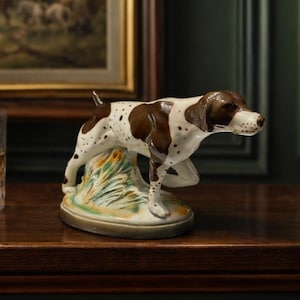 Puede incluir: Una figura de cerámica de un perro Pointer, con marcas marrones y blancas, posada sobre una base herbosa. El perro está colocado sobre una superficie de madera oscura, con una pintura enmarcada y una botella de whisky en el fondo. También se ve un vaso de whisky.
