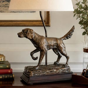May include: A table lamp featuring a bronze-toned sculpture of a standing hunting dog as the base. The lamp has a simple, tapered off-white fabric lampshade and a dark rectangular base. The dog is depicted in a mid-stride pose with its tail raised.