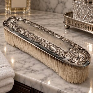 May include: An oval-shaped clothes brush featuring a polished silver-tone handle with intricate embossed scrollwork and floral detailing along the edges, paired with dense, light-colored natural bristles for garment maintenance.