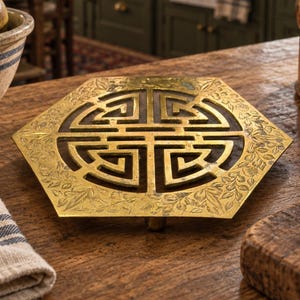 May include: A hexagonal brass trivet featuring an intricate openwork geometric pattern in the center and a detailed floral engraving along the outer border. The metal surface has a polished finish with visible signs of aged patina and texture.