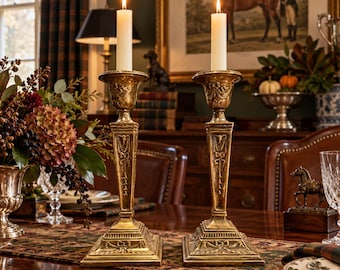 May include: A pair of matching antique-style gold-toned metal candlesticks with square bases and intricate relief detailing, each holding a single lit white taper candle, positioned on a dark wooden dining table surface.
