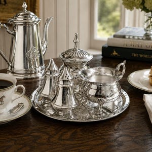 May include: A silver tea service set including a tall teapot, a lidded sugar bowl, a creamer, and two conical shakers arranged on a round silver tray. The pieces feature polished metallic surfaces and ornate detailing, reflecting light on a dark wooden surface.