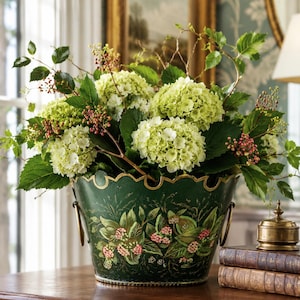 May include: A dark green metal bucket with a scalloped rim and gold-painted edges, featuring hand-painted floral motifs of green leaves and pink berries. The bucket has two side handles and is filled with a large arrangement of green hydrangeas.