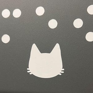 May include: A white cat face silhouette with whiskers and ears, set against a dark gray background. Above the cat's head are several white circular shapes, resembling bubbles or stars. The design is simple and graphic.