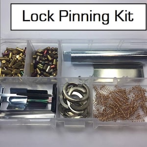 May include: A clear plastic case containing a selection of lock picking tools. The case has six compartments, each holding different types of tools, including pins, springs, and a metal rod. The case is labelled "Lock Pinning Kit".