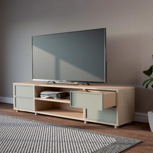 May include: A light wood TV unit with two light green drawers and an open shelf. The unit has a contemporary design. A large flat-screen TV is on top, and a rug is in front. The unit is 150cm wide.