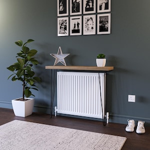 May include: A white radiator with black hairpin legs sits on a wooden shelf. The shelf is mounted on a wall with a dark green paint colour. A potted plant and a star-shaped decoration are on the shelf.