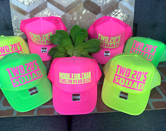 May include: Five neon green and pink trucker hats with mesh backs. The hats have the text "TWO 20'S SQUAD" in pink lettering on the front. One hat has the text "MORE FUN THAN TWO 20 YEAR OLDS" in yellow lettering.