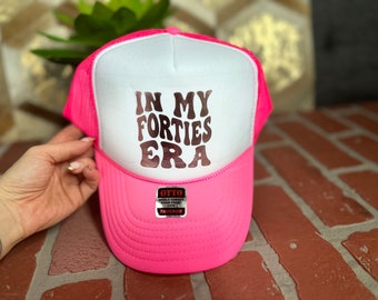 Puede incluir: Gorra de camionero rosa y blanca con el texto "In My Forties Era" en una fuente de lámina dorada.