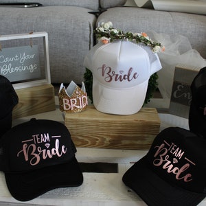 May include: Five black and white trucker hats with the words "Team Bride" in pink script. One white trucker hat with the word "Bride" in pink script. A gold glitter crown with the word "Bride" on it.