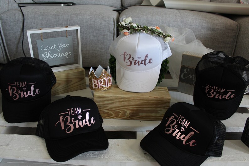 May include: Five black and white trucker hats with the words "Team Bride" in pink script. One white trucker hat with the word "Bride" in pink script. A gold glitter crown with the word "Bride" on it.