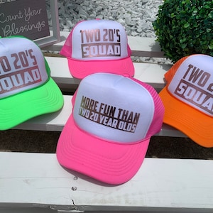 May include: Four trucker hats with a white front panel and a mesh back. The hats have different colored brims: green, pink, orange, and pink. The front panel of each hat has the text "TWO 20'S SQUAD" in rose gold lettering. One hat has the additional text "MORE FUN THAN TWO 20 YEAR OLDS" in rose gold lettering.